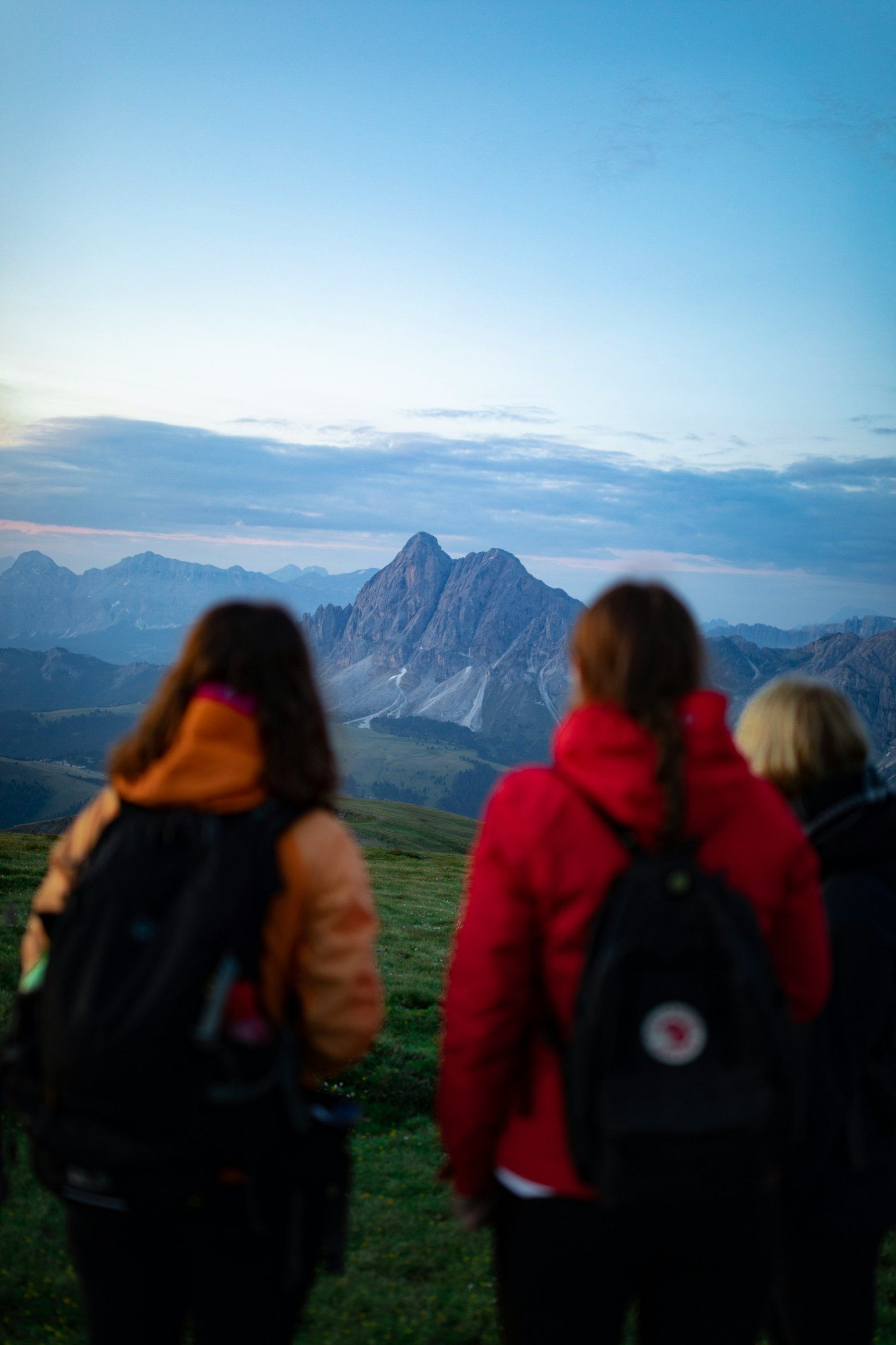 Three women in warm jackets look out across a green ridge toward a jagged mountain peak at dawn.